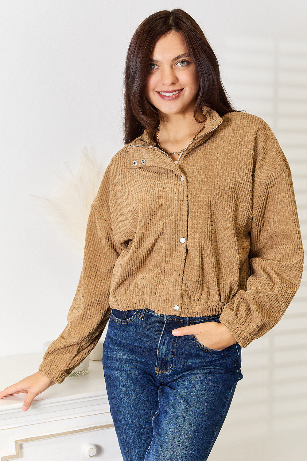 Camel cropped jacket with a raised grid texture, stand collar and snap placket over a front zip, worn closed with blue jeans, front view