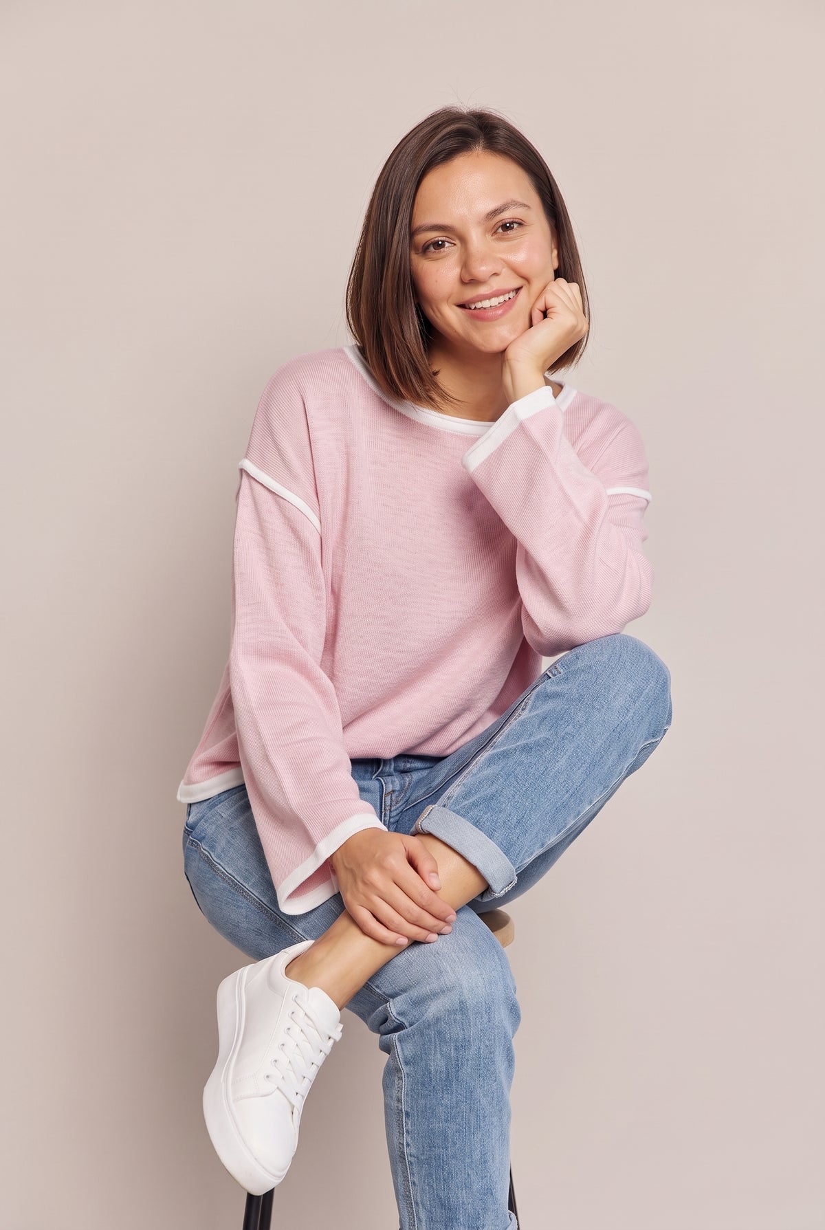 Seated view of the blush pink boat neck knit top with white trim, chin resting on one hand