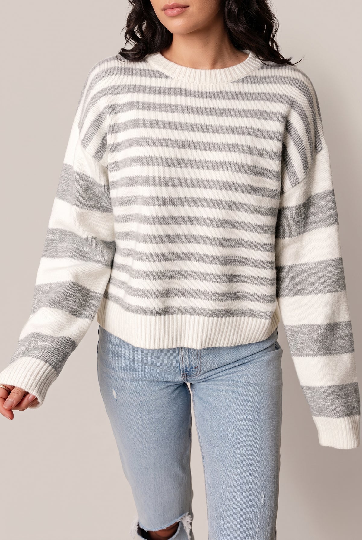 Grey and off-white striped crew neck sweater with dropped shoulders and wide-striped long sleeves, worn with light wash jeans, front view