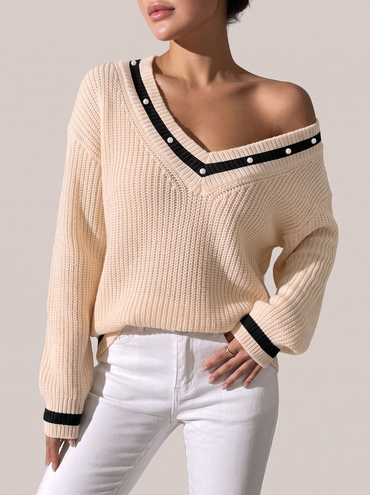 Tan V-neck sweater worn off one shoulder, with black trim and pearl-look beads along the neckline and black-striped cuffs